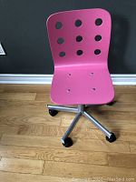Front view of pink IKEA children's desk chair with backrest and caster wheels on wooden floor against dark wall.