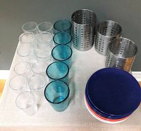 Set of 11 clear ribbed glasses, 5 blue plastic tumblers, 7 small plates in blue, red, and white, and 3 perforated stainless steel metal canisters arranged on a pale surface