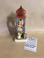 Front view of the angel bell tower figurine showing three angel figures, clock face, and reddish-orange dome top.