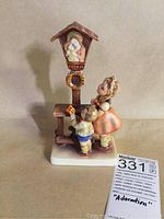 Front view of M.J. Hummel porcelain figurine showing two children bringing flowers to a religious shrine icon, placed on a white square base with visible seller's label