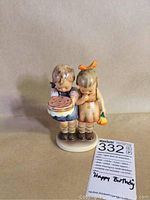 Front view of M.J. Hummel figurine showing two children holding a cake, with auction tag visible labeled 'Happy Birthday'.