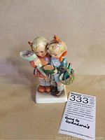 Front view of porcelain figurine showing two children holding a bowl, flowers, and a basket with items. The figurine is on a rectangular base with handwritten label.