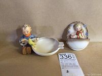 Photo showing two Hummel figurines. Left: boy with lute and bird attached to ash receiver dish. Right: angel bird holy water font with decorative details.