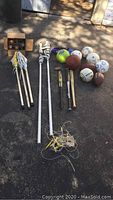 Overview showing volleyball net poles, lacrosse sticks, various balls including basketball, baseballs, footballs, soccer balls, kickball, and baseball bats, and a wooden crate with more items