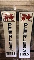 Two vertical white metal Peerless Tires signs with red griffin logos and black text standing against a wooden wall.