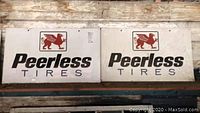 Two white metal Peerless Tires signs side by side on a wooden surface showing full design and size.