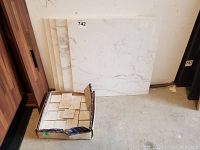 Four large square marble tiles leaning against a wall with a box of small tiles in front on the floor.