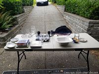 Table set outdoors on driveway with white stoneware dish, wine glasses, decorative glass items, cookbooks, and metal drying rack visible.