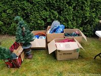 Outdoor photo showing a small artificial Christmas tree in a urn, multiple cardboard boxes with Christmas decorations and lights, and a folding table.