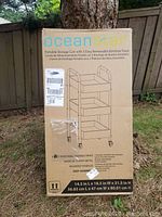Box of Ocean Star portable storage cart with 3 removable bamboo trays shown outdoors against a tree and wooden fence.