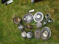 Overview photo showing the collection of silver plated items spread out on grass, including cups, trays, covered dishes, and a vintage cigar holder.