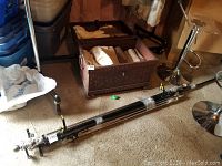 Three bundled adjustable black metal curtain rods with curtain rings and light curtains on the floor alongside a metal chest (not included).