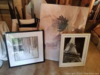 Three framed art prints in a storage or warehouse setting. Visible prints include an abstract large print with vertical lines, a black and white snowy forest scene print in a dark frame, and a black and white Eiffel Tower photograph in a white frame.