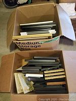 Two open cardboard boxes each filled with assorted size picture frames in black, white, gold-tone colors.