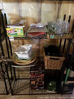 Metal shelf with assorted kitchen items including plates, bowls, glasses, basket with utensils, and boxed items below