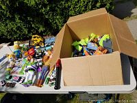 Wide view of assortment of toys on white table including plush toys, toy vehicles, figures, and a cardboard box with toy track pieces inside