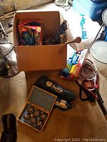 Photo showing cardboard box with tennis balls, racket handles, and various outdoor game items, and wooden box with bocce balls and target ball.