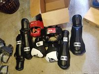 Photo of sparring gear including shin guards, gloves, and red headgear arranged on floor near cardboard box.