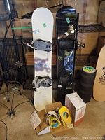 Two snowboards standing vertically side by side with bindings attached; a white Atlantis 155cm and a black Solomon 153cm, plus extra snowboard bindings in a box in front.