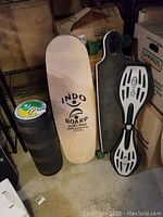 Photo showing Indo Board roller, balance board wooden board, and two longboards against wall with boxes.