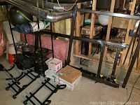 Wide shot of wire shelving with stacked weight plates, fixed hex dumbbells, adjustable dumbbells, and disassembled squat rack frame leaning behind shelving.