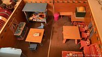 Top-down view of the dollhouse interior showing bed with doll, multiple tables, chairs, cupboards, and painted floors and walls.