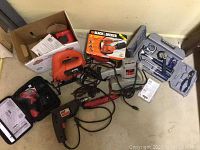 Photo showing grouped power tools including jigsaw, Black & Decker sander in box, various hand tools in plastic case, and drill.