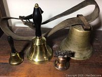 Five bells displayed on a wooden surface; includes one large cow bell with leather strap and four smaller hand bells of varying sizes and metal tones