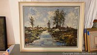 Framed oil painting showing a landscape with water, trees, houses, and windmills.