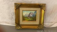 Framed small mountain landscape painting hanging against fabric background.