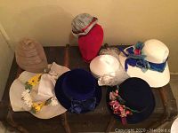 Seven vintage hats arranged on a flat surface along with a red mannequin head covered in fabric. Hats include various colors and styles with ribbon, bow, and flower detailing.