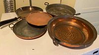 Overall view of vintage copper cookware including pans and colander on table.