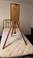 Full view of wooden artist easel standing on a mattress with framed cartoon artwork attached