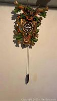 Full view of the wood carved wall clock hanging on a wall showing the clock face, weight, carved deer head, rifles and oak leaves.