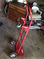 Red metal hand truck dolly with two large black rubber wheels and two smaller caster wheels, positioned in a garage with various furniture and items in background.
