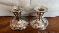 Pair of two weighted sterling silver candlesticks on wooden surface with white cloth backdrop, showing the front and side angles along with decorative base and stem details.