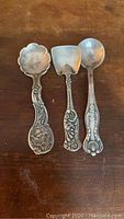 Three sterling silver salt spoons placed side-by-side on a wooden surface, showing the front view and detail of each spoon's unique handle and bowl shape.