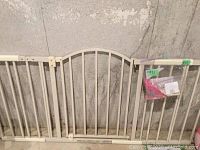 Two white metal baby gates leaned against a concrete wall, one gate has a slightly arched top. A pink plastic bag with small hardware pieces hangs from one gate.