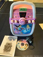 Front view of Disney Princess themed karaoke machine showing control panel and character image, with Party Tyme CD, power adapter, and owner's manual in front