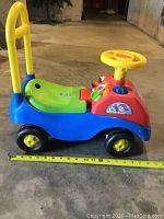 Side view of plastic ride-on push toy showing yellow back handle, green seat, red front panel with yellow steering wheel and four wheels. Measuring tape indicates about 22 inches long.