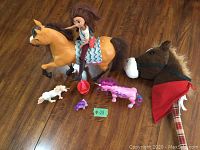 Wide shot of various horse and animal toys on wooden floor including large horse with doll rider, plush hobby horse, small figures, and pink unicorn