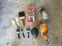 Paint brushes, mini roller, paint sprayer, hand sander, and electric sander arranged on concrete floor.