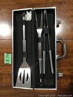 Three stainless steel BBQ utensils—flipper spatula, tongs, and two-pronged fork—secured inside a silver metal case during open display.