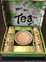 Six small tea cups with matching saucers in green and white tea-themed box. Cups and saucers cream-colored with varied "Tea" text designs and chair illustrations in black and green.