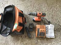 Ridgid drill set including drill, battery charger, non-working battery, side handle, manual, and carrying bag laid out on carpet