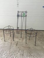 Three black metal flower pot holders in varying heights displayed on concrete floor against white wall.