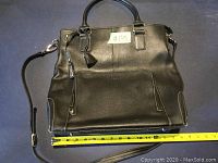 Black L. Credi leather handbag shown from front, displaying zippered pocket with tassel pull and top handles. Measuring tape shows approximate width of 14 inches.