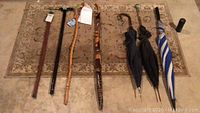 Photo showing four walking canes, three umbrellas, and a small rug underneath all items on the floor