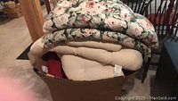 Photo of large floral patterned cushions stacked on top of beige and solid cushions inside a cardboard box.