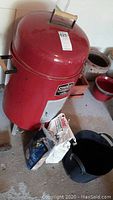 Red cylindrical Char-broil water smoker with front access door and domed lid with handle, sitting on a concrete floor with charcoal bags and bucket near it.